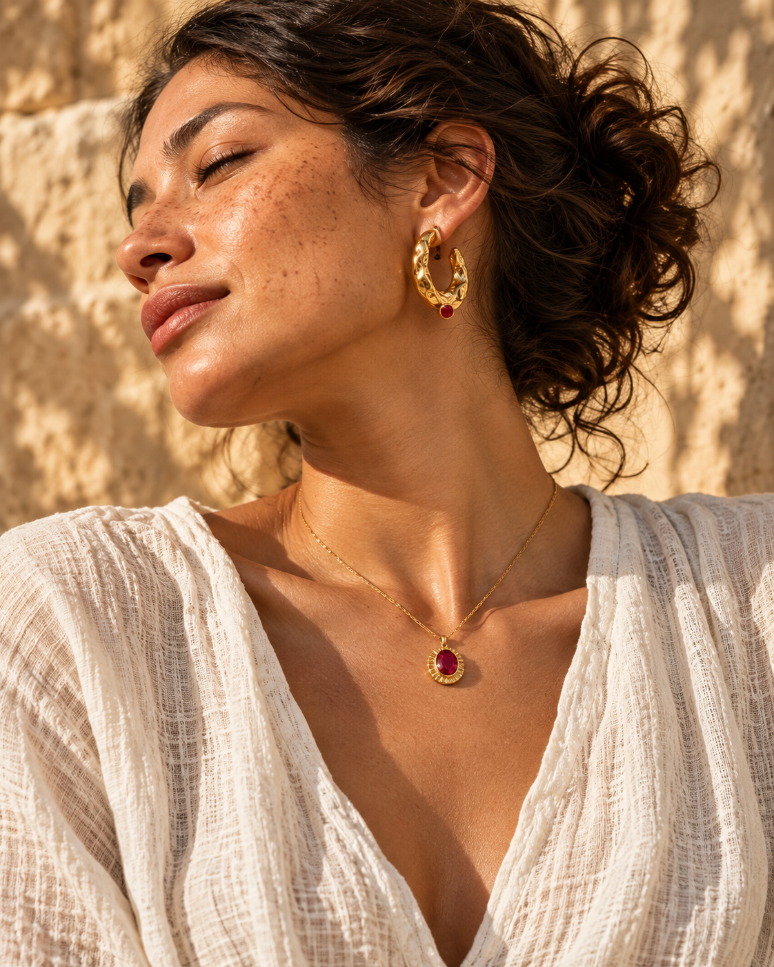 Woman in white linen wearing a gold hoop and ruby pendant in warm sunlight